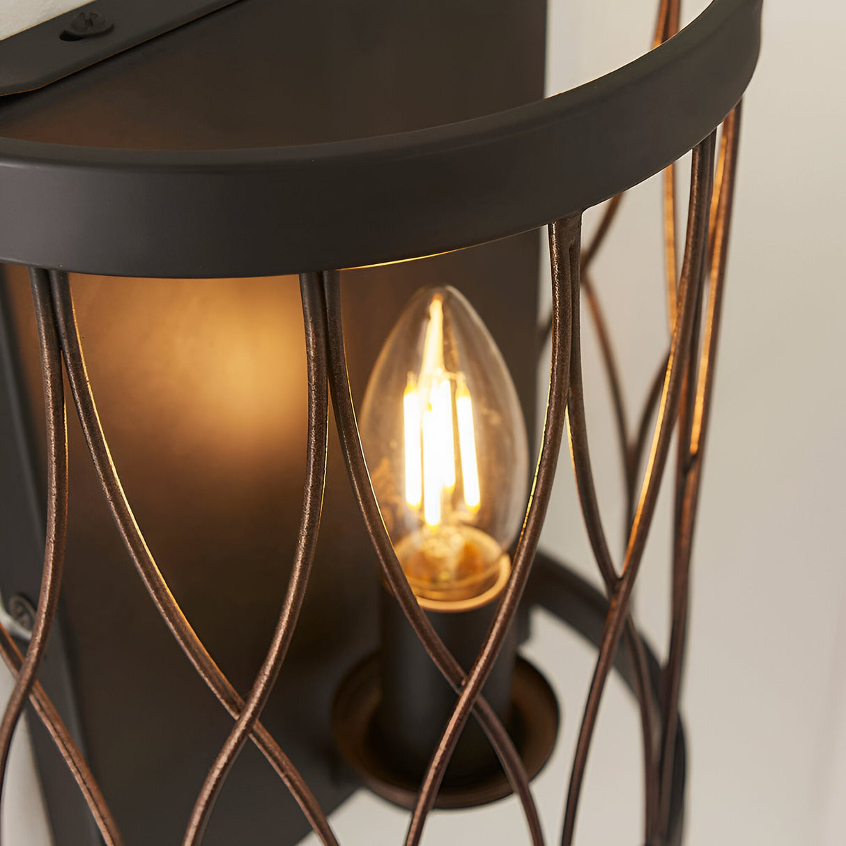 Close-up of Endon Hulton wall light in matt black and rustic bronze, showing the lit E14 candle bulb within the curved steel cage frame.