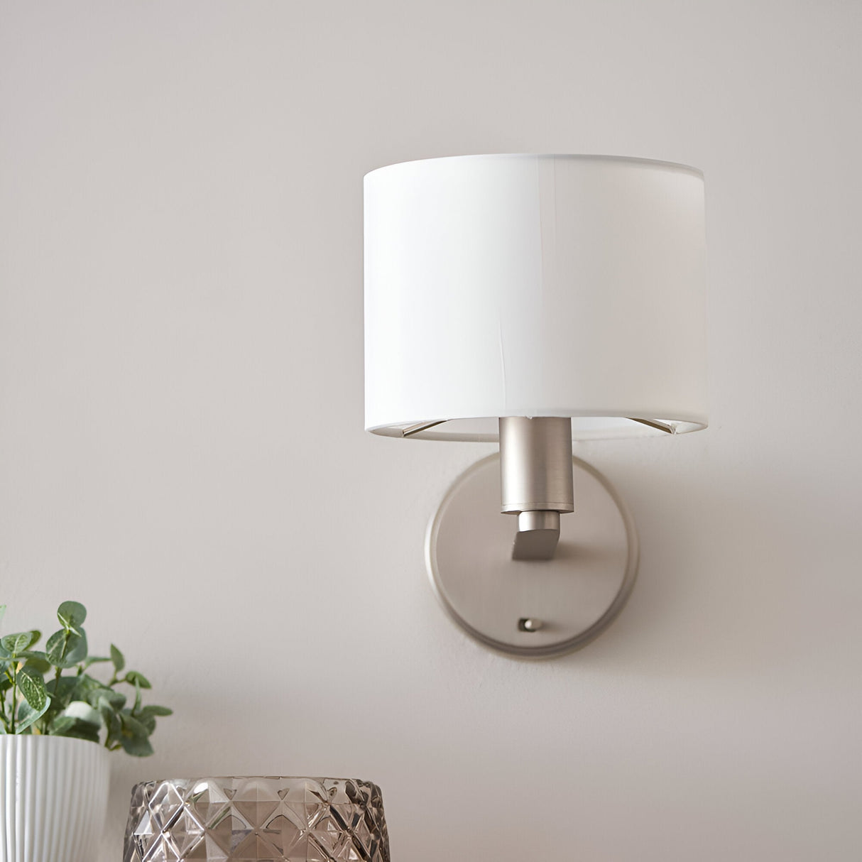 Bexley Wall Light in nickel with a white faux silk drum shade, shown mounted on a light grey wall above a plant and glass decor.