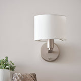 Bexley Wall Light in nickel with a white faux silk drum shade, shown mounted on a light grey wall above a plant and glass decor.
