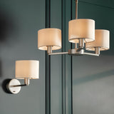 Bexley Wall Light in nickel with white drum shade, mounted on a dark green panelled wall alongside a matching chandelier.