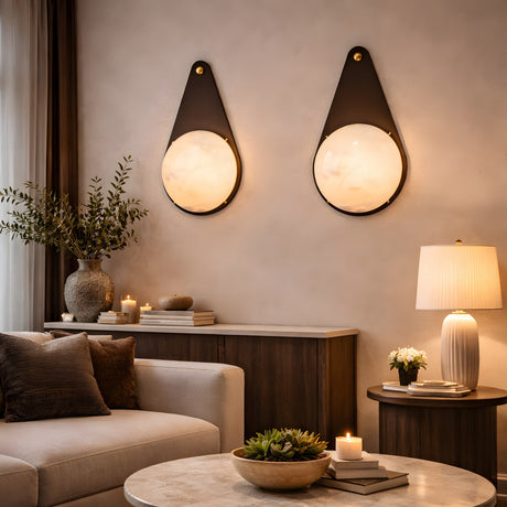 A cozy living room with a beige sofa, wooden furniture, potted plants, candles, and two Lunara 1 Light Teardrop Alabaster Wall Lights in Matte Black, casting an elegant circular glow for a warm, inviting atmosphere.
