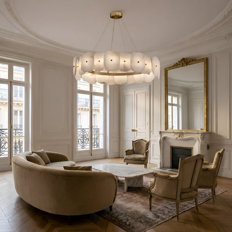 Elegant living room with cream walls, large windows, a gold-framed mirror above a white fireplace, marble coffee table, tan curved sofa, two armchairs, and the Solvane LED Medium Petal Alabaster Chandelier in Soft White & Brass.