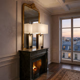 Elegant room with a marble fireplace, lit fire, and the Valcera 1 Light Framed Vertical Alabaster Table Lamp in Black & Soft White beside a large mirror. Tall windows showcase a sunset cityscape, while warm lighting adds cozy charm.