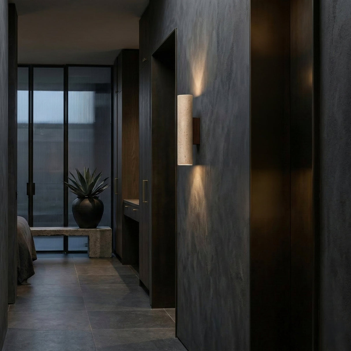 A modern hallway with walnut brown walls and floors features the Dorsa 1 Light Large Cylinder Travertine Wall Light in Walnut Brown & Light Beige. A large potted plant sits by floor-to-ceiling windows; a bedroom is partially visible to the left.