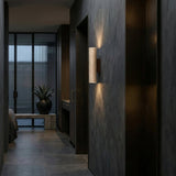 A modern hallway with walnut brown walls and floors features the Dorsa 1 Light Large Cylinder Travertine Wall Light in Walnut Brown & Light Beige. A large potted plant sits by floor-to-ceiling windows; a bedroom is partially visible to the left.