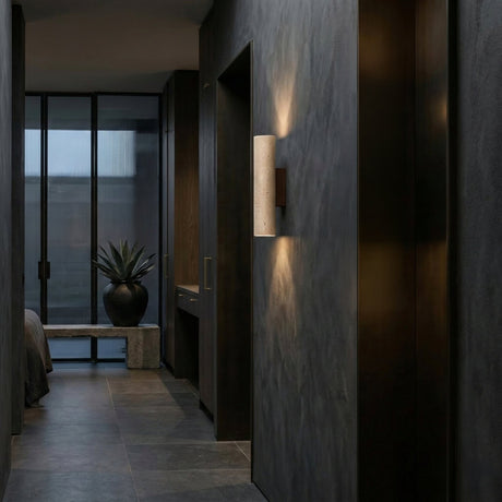 A modern hallway with walnut brown walls and floors features the Dorsa 1 Light Large Cylinder Travertine Wall Light in Walnut Brown & Light Beige. A large potted plant sits by floor-to-ceiling windows; a bedroom is partially visible to the left.