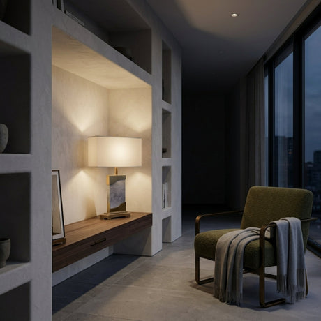 A modern reading nook with a green armchair, gray blanket, wooden built-in shelf, and the Torvani 1 Light Statement Marble Table Lamp in brass, grey & white. Framed art decorates the dim room with large windows and city views.