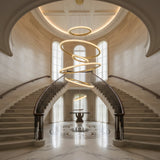 A grand foyer with twin curved staircases, a geometric marble floor, tall arched windows, and the Lucenza LED Cascading Halo Ring Alabaster Chandelier in brushed brass & soft white illuminating a round table. The space is bright, elegant, and symmetrical.