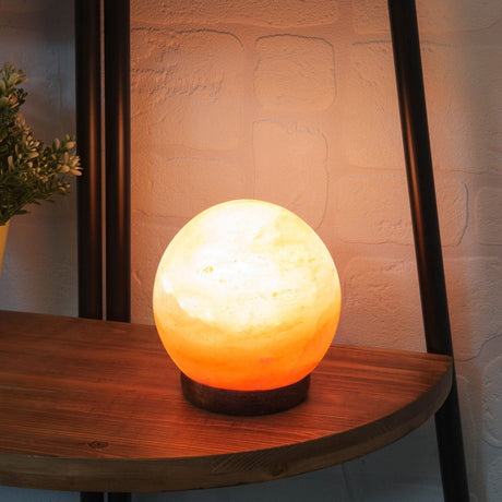 Sphere Himalayan rock salt lamp, 13.5cm, glowing orange and cream on a round dark wood base, displayed on a wooden shelf.