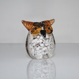Miniature glass owl figurine with round white-speckled clear glass body, brown head, pointed ear tufts, and wide brown eyes, set against a plain light background.