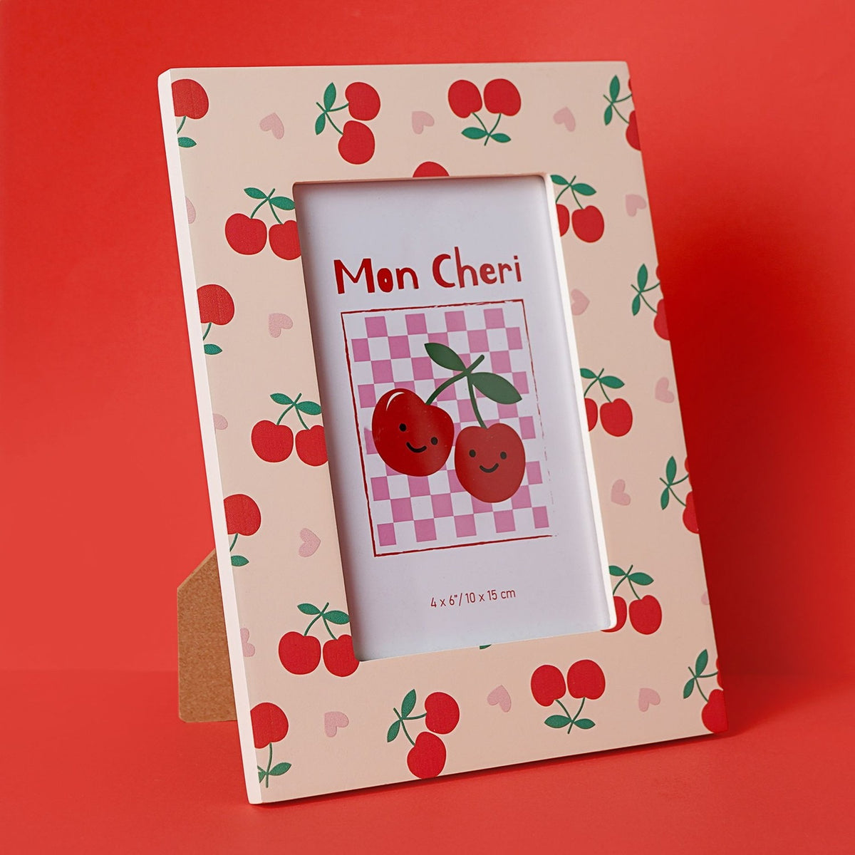 Wooden rectangular photo frame with stand in peach, printed with red cherries, green stalks and pink hearts, displaying a "Mon Cheri" cherry graphic insert, against a red background.