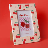 Wooden rectangular photo frame with stand in peach, printed with red cherries, green stalks and pink hearts, displaying a "Mon Cheri" cherry graphic insert, against a red background.