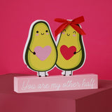 Wooden plaque featuring two smiling avocado figures holding pink and red hearts, one wearing a red ribbon bow, set on a pale pink base reading "You are my other half".