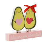 Wooden plaque with two smiling avocado characters holding pink and red hearts, one wearing a red ribbon bow, on a pale pink base reading You are my other half.