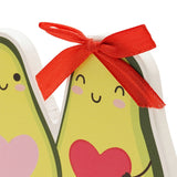 Close-up of the Other Half Avocado Couple wooden plaque, showing smiling green avocado faces with rosy cheeks, pink and red hearts, and a red ribbon bow on white backing.