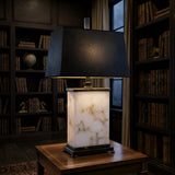 The Noctaryn 1 Light Rectangular Alabaster Table Lamp in Black & Soft White sits on a wooden table in a dimly lit library, surrounded by bookshelves and a window showing the evening sky.