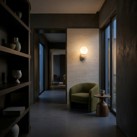 A modern hallway with dark walls, a green armchair, a round wood side table, shelves with pottery, and the Belaro 1 Light Globe Travertine Wall Light in light beige adding warmth as large windows fill the space with natural light.