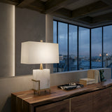 The Lumenis 1 Light Alabaster Table Lamp in Brass & Soft White sits on a wooden sideboard near large windows with city views, alongside books and decor, adding an elegant architectural lighting touch to the space.