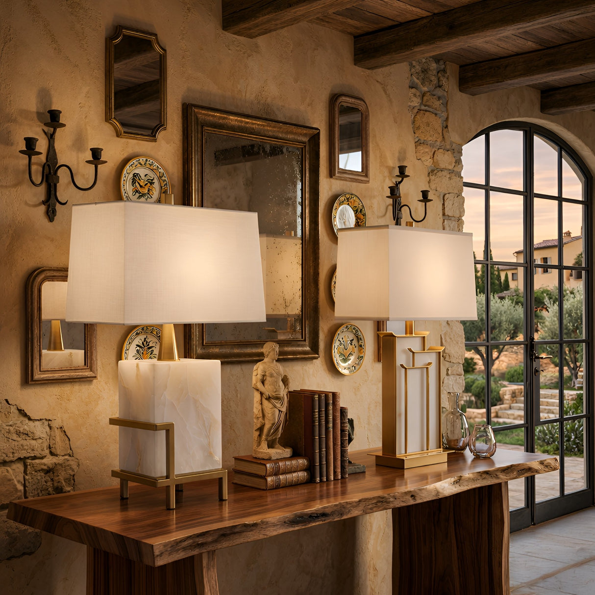 Elegant rustic room with a wooden table, books, decorative plates, a small statue, framed mirrors on a stone wall, and the Veliora 1 Light Framed Alabaster Table Lamp in brass & soft white; large window reveals a scenic sunset view.