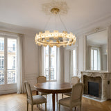 A round wooden dining table with four beige chairs sits in an elegant room with large windows, a decorative fireplace, a large mirror, and the Ravion LED Small Panelled Alabaster Chandelier - Soft White & Brass hanging from the ceiling.