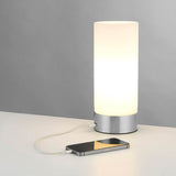 Endon Bracewell nickel table lamp with brushed metal base, matt opal glass cylindrical shade and USB port, shown charging a smartphone on a light surface.