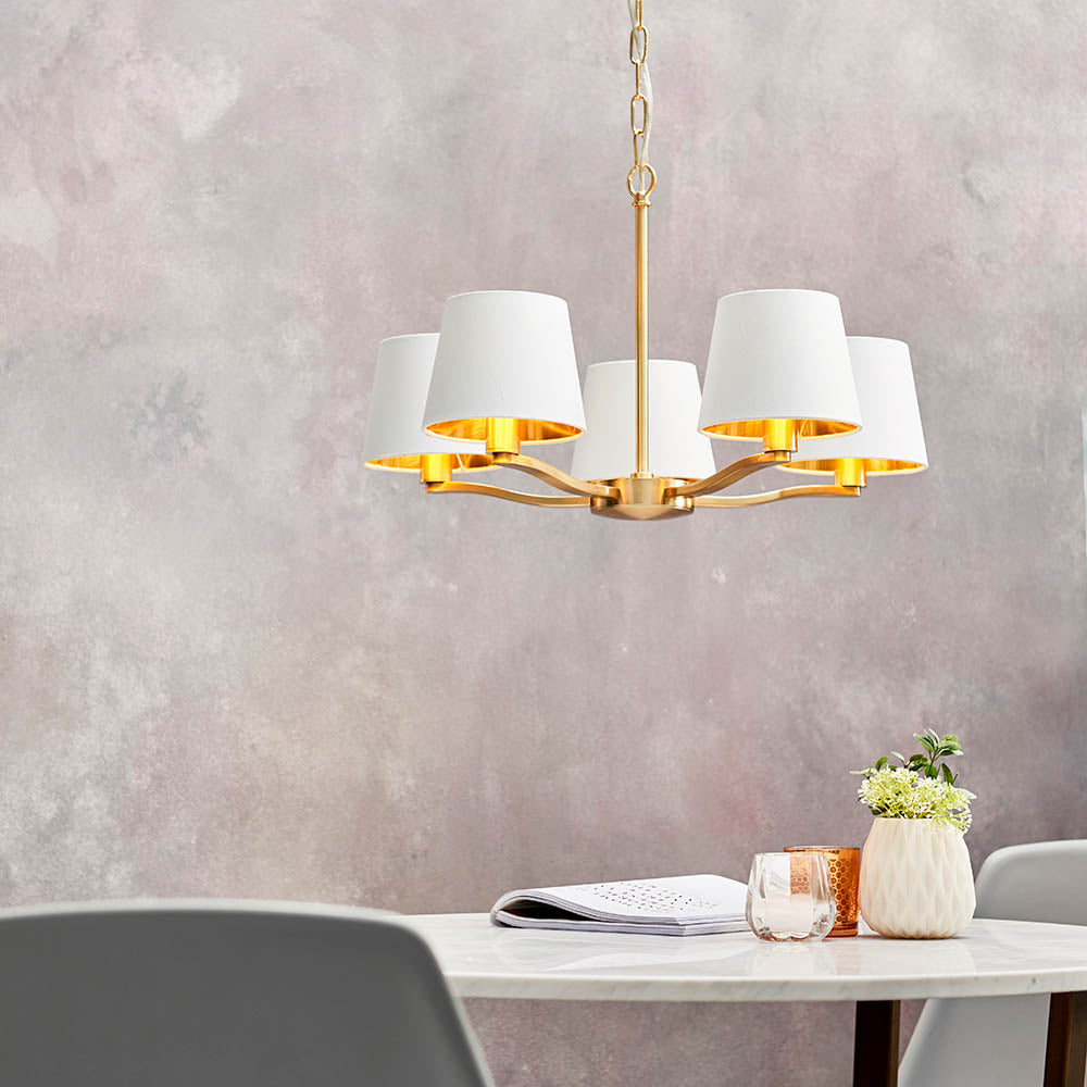 Brendale 5 Light Pendant in satin gold, with white faux silk shades and gold inner linings, hung above a dining table in a soft grey room.