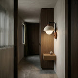 A minimalist hallway with wood-paneled walls, a floating wooden shelf with a vase, and the Corvella 1 Light Dome Travertine Wall Light in Walnut Brown & Light Beige above, plus a window with a green curtain that bathes the space in ambient light.