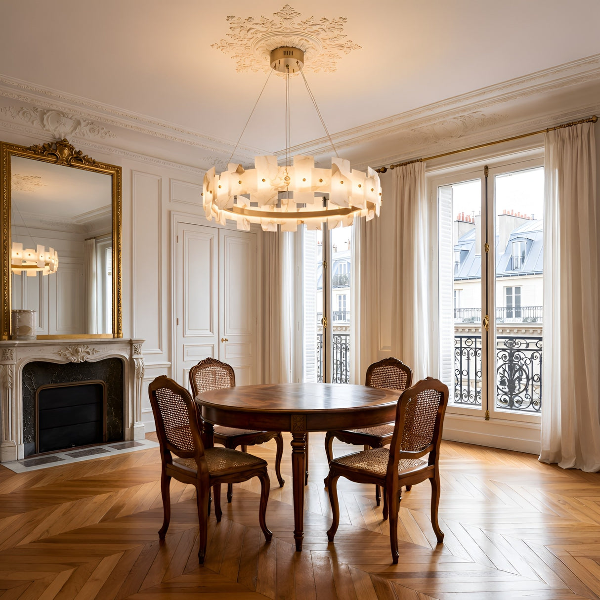Elegant dining room with a round wooden table, four vintage chairs, a decorative fireplace with a large mirror, parquet floors, tall windows with sheer curtains, and the Ravion LED Medium Panelled Alabaster Chandelier in soft white & brass overhead.