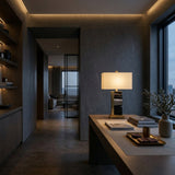 A modern study with a sleek wooden desk, the Vellum 1 Light Structured Form Marble Table Lamp in brass and black, books, and decor by a large window. Built-in shelves and a hallway to the living area are seen in the background.