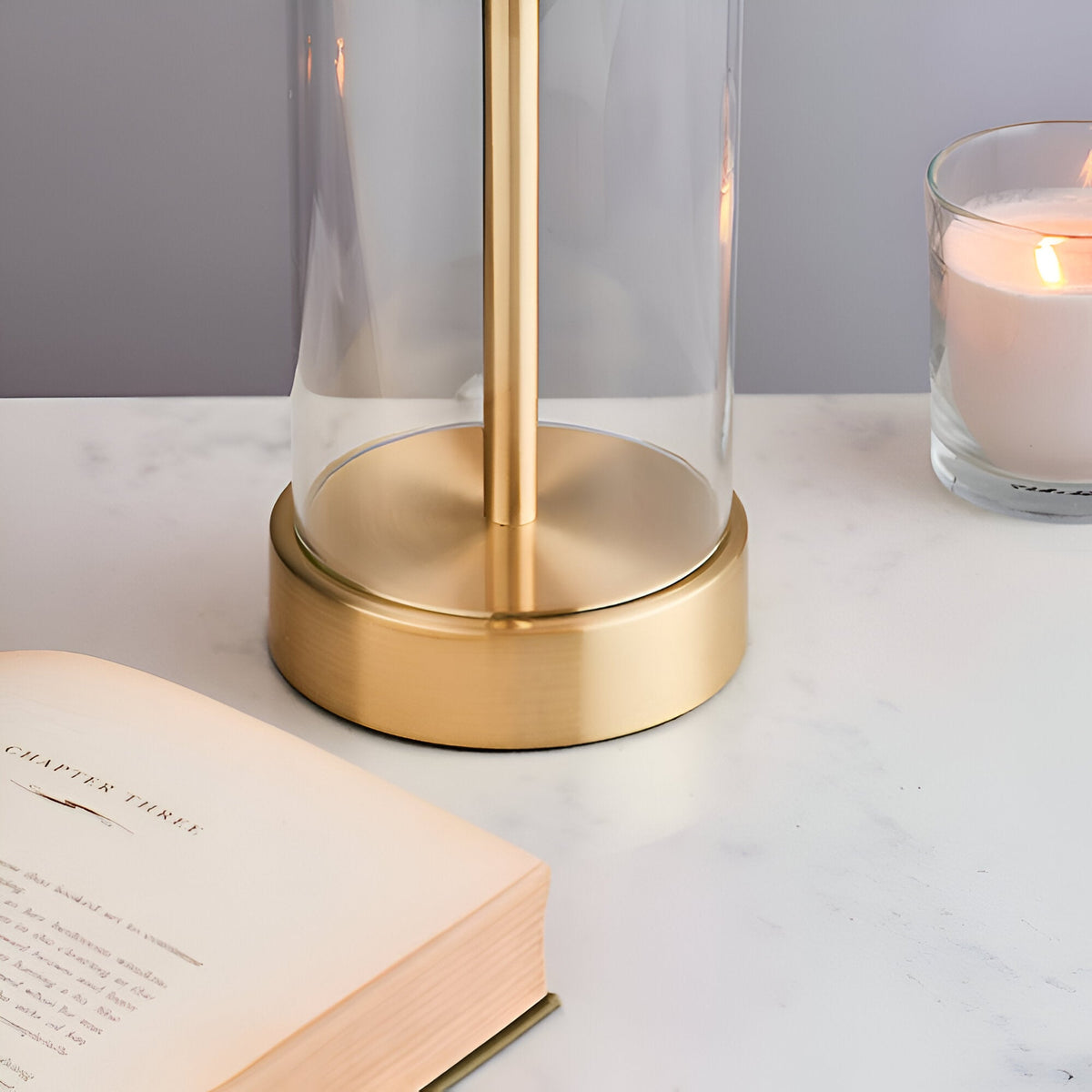 Endon Caldley table lamp base in satin brass with clear cylindrical glass, shown on a marble surface beside an open book and lit candle.