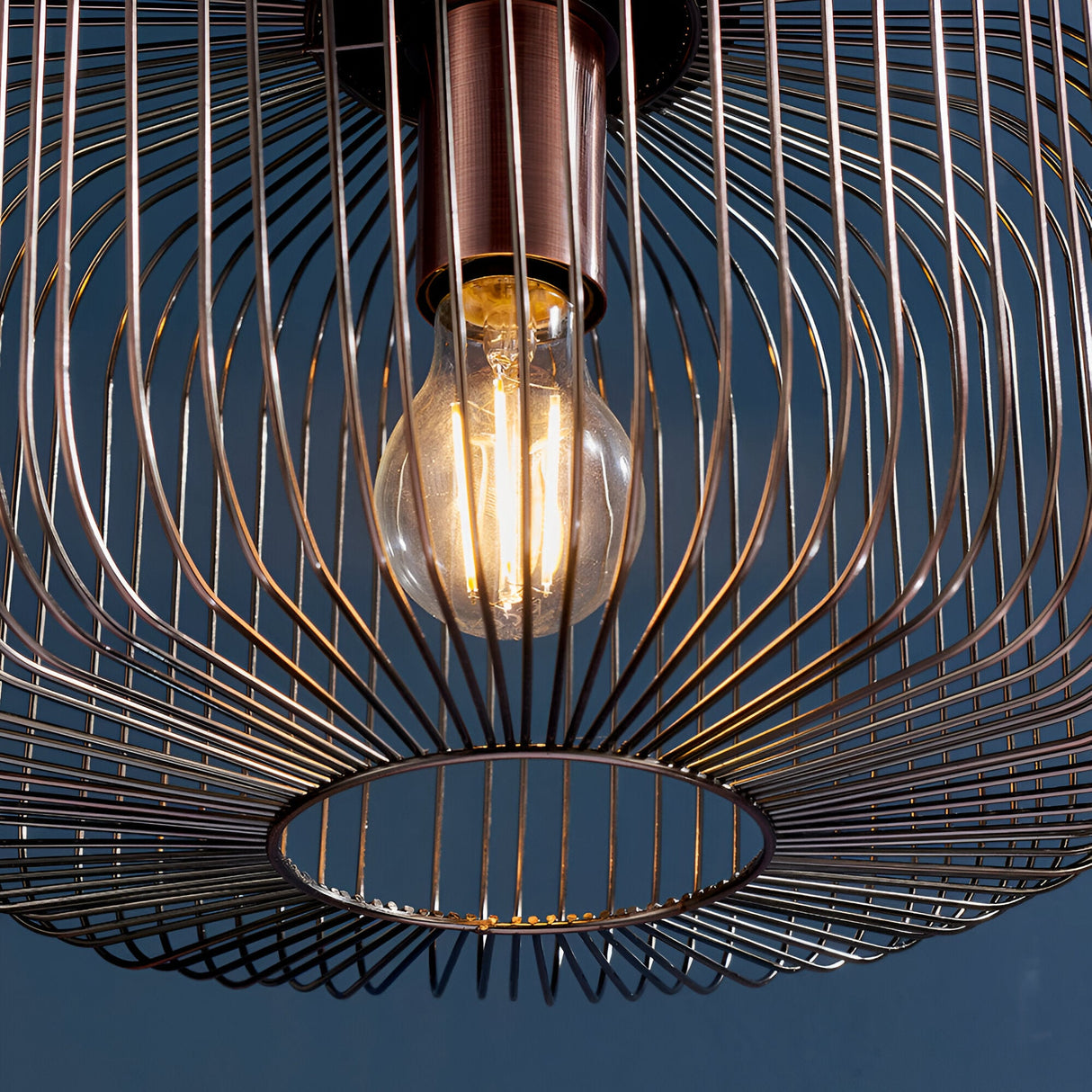 Castdale Cage Pendant in antique copper, close-up of the open wire cage shade with visible E27 bulb, set against a dark blue background.
