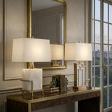 A stylish console table with books, a brass sculpture, and the Lumenis 1 Light Alabaster Table Lamp – Brass & Soft White sits before a large mirror and window in an elegant beige room with gold accents.