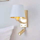 Brendale Wall Light in satin gold with a white fabric shade and adjustable LED reading spot, shown mounted above a bed in a bedroom setting.