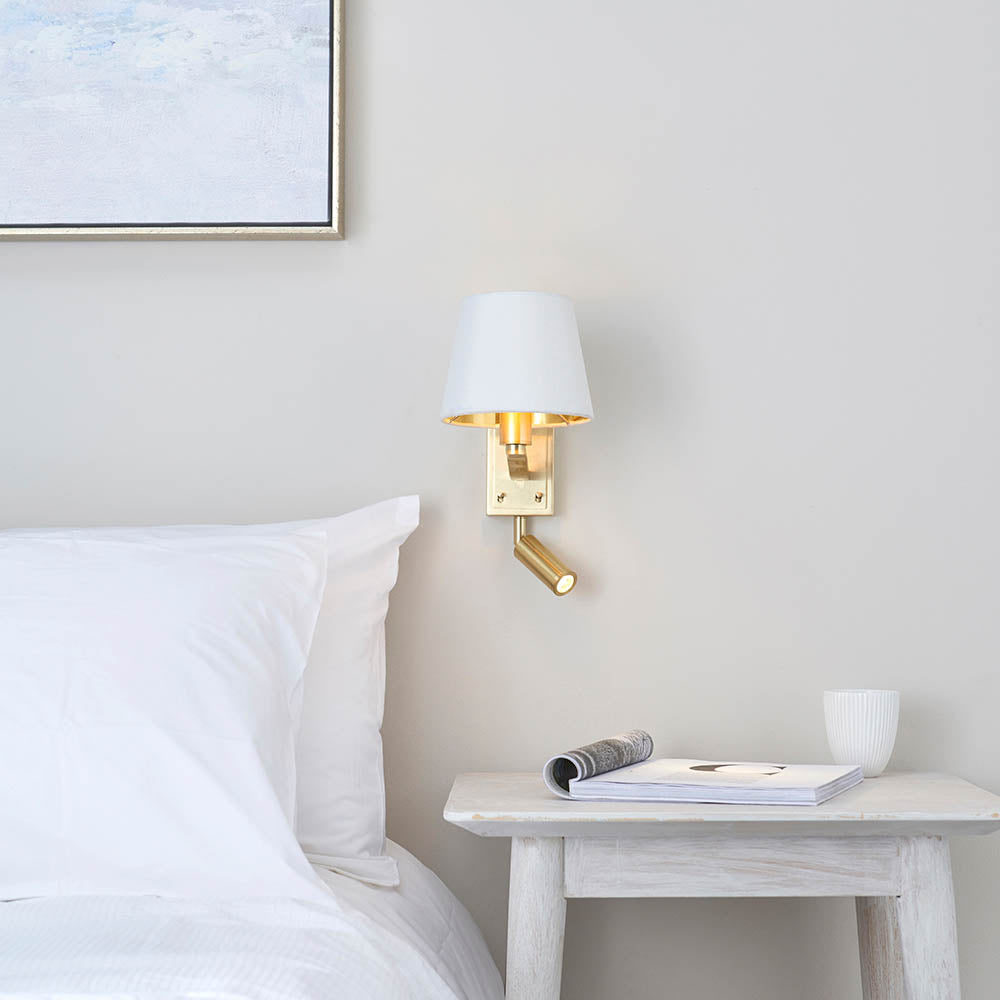 Brendale Wall Light in satin gold with a white shade and adjustable LED reading spot, shown above a bedside table in a bedroom setting.