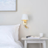 Brendale Wall Light in satin gold with a white shade and adjustable LED reading spot, shown above a bedside table in a bedroom setting.