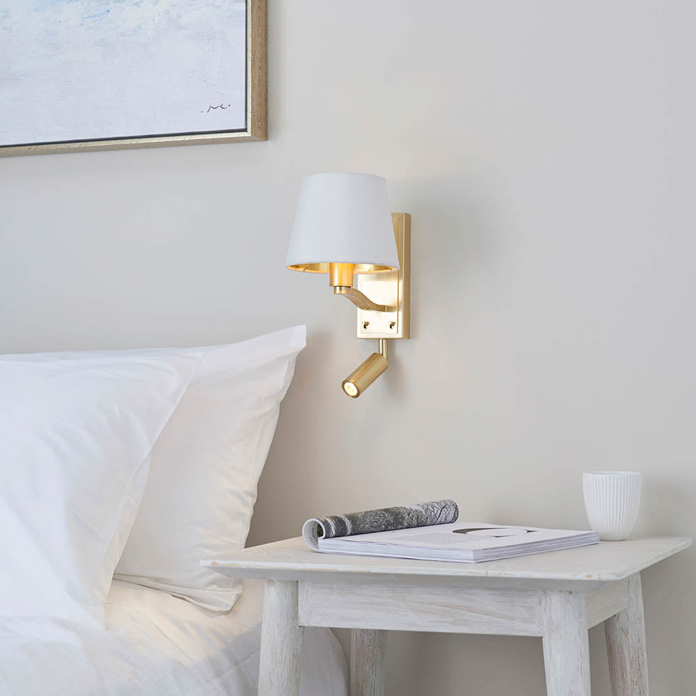 Brendale Wall Light in satin gold with white faux silk shade and adjustable LED reading spot, shown above a bedside table in a bedroom setting.