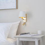 Brendale Wall Light in satin gold with white faux silk shade and adjustable LED reading spot, shown above a bedside table in a bedroom setting.