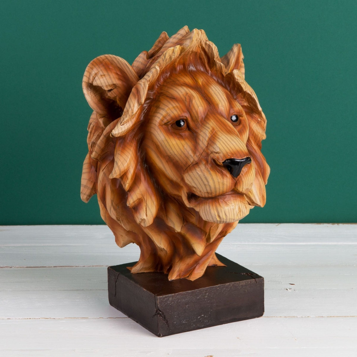 Lion head figurine in brown wood-effect resin with layered mane, glossy eyes and black nose, mounted on a square dark brown base against a green background.