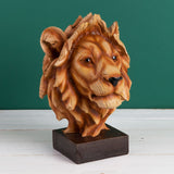 Lion head figurine in brown wood-effect resin with layered mane, glossy eyes and black nose, mounted on a square dark brown base against a green background.