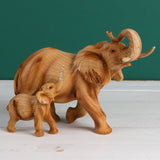 Elephant and calf wood effect resin figurine in brown, both trunks raised, with visible wood grain detailing and cream tusks, set against a dark green background.