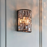 Endon Aldwick wall light in dark bronze finish with tiered faceted clear crystals, single E14 lamp illuminated, mounted on a pale panelled wall.