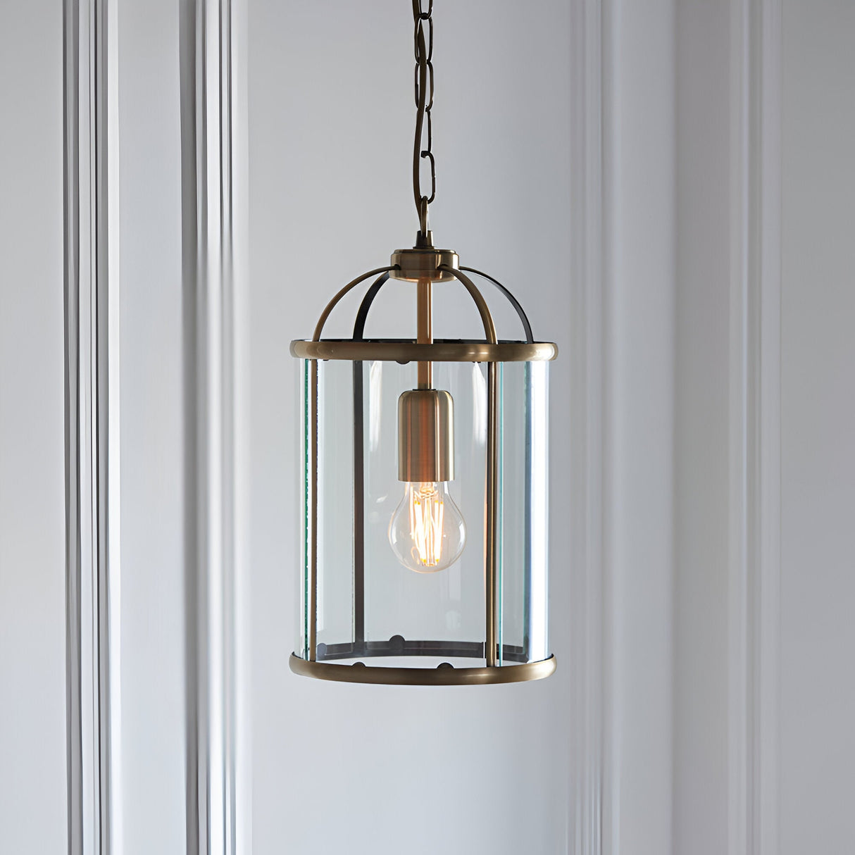 Caldbury Pendant Light in brass, cylindrical lantern frame with clear glass panels and exposed E27 bulb, hung from a chain against a plain white wall.