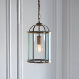 Caldbury Pendant Light in brass, cylindrical lantern frame with clear glass panels and exposed E27 bulb, hung from a chain against a plain white wall.