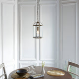 Caldbury Pendant Light in brass, single E27 lantern-style fixture with clear glass panels, hung above a dining table in a panelled white room.