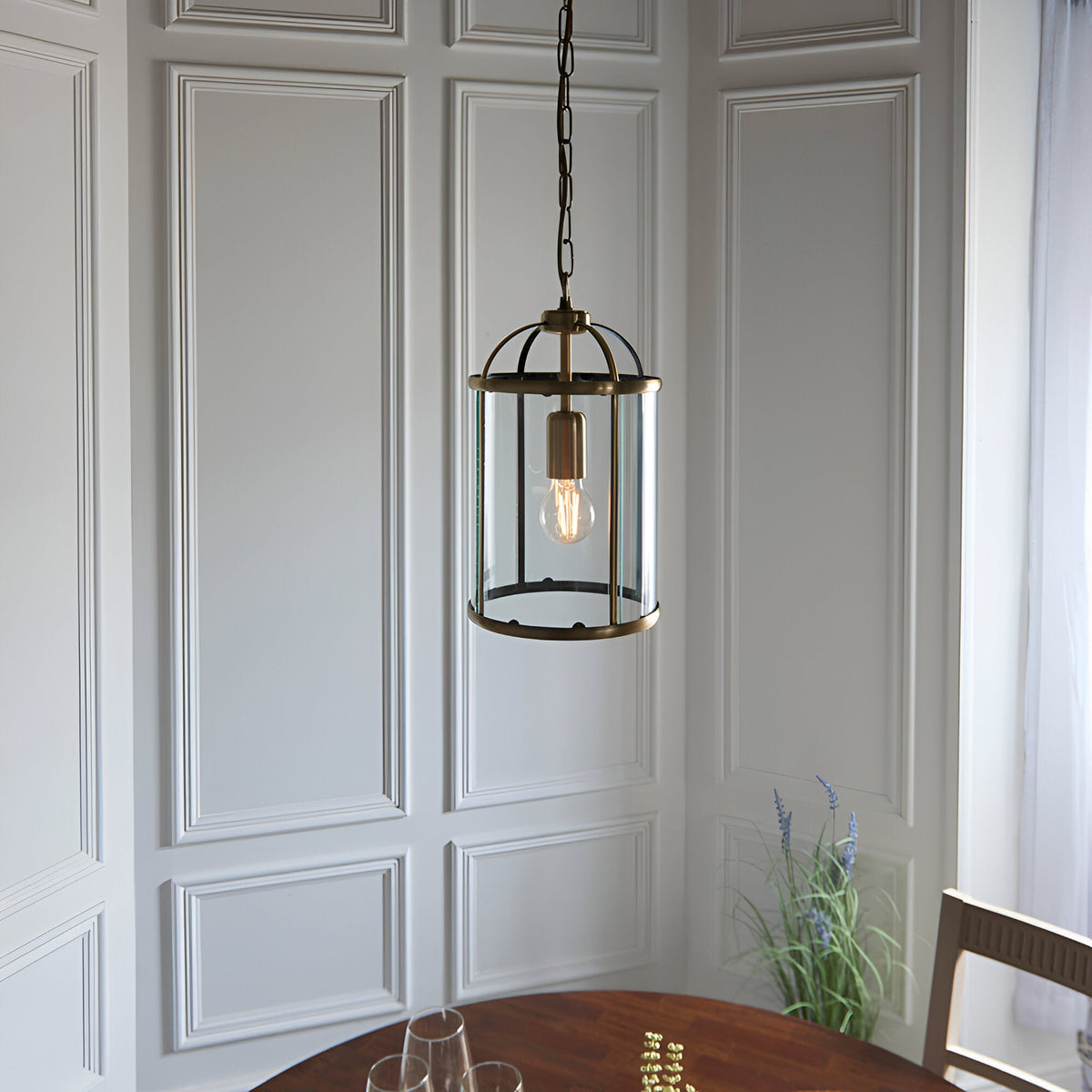 Caldbury Pendant Light in brass, a lantern-style fixture with clear glass panels and exposed E27 bulb, hung from a chain above a dining table.