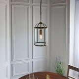Caldbury Pendant Light in brass, a lantern-style fixture with clear glass panels and exposed E27 bulb, hung from a chain above a dining table.