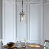 Caldbury Pendant Light in brass, a single lantern-style fixture with clear glass panels and exposed bulb, hung from a chain above a dining table.