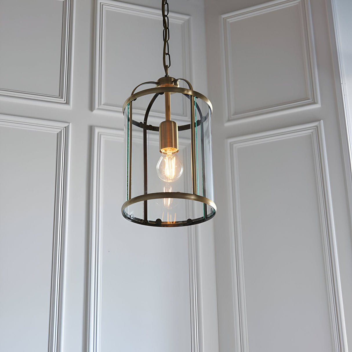 Caldbury Pendant Light in brass, a cylindrical lantern-style fixture with clear glass panels and a visible filament bulb, hung from a chain.