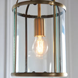 Caldbury Pendant Light in brass, cylindrical clear glass panel lantern frame with an exposed E27 filament bulb, shown close-up against a light background.