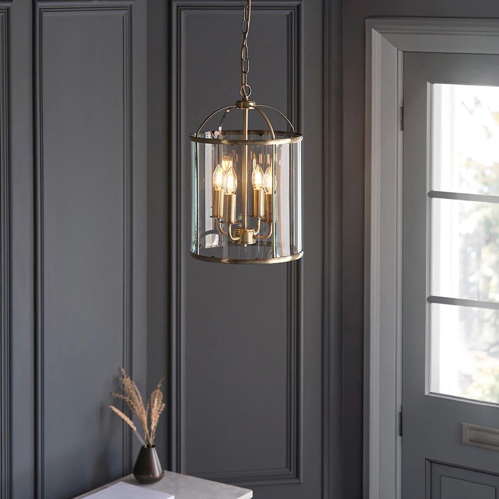 Caldbury 4 Light Pendant in brass with clear glass panels and candle lamps, hanging from a chain in a dark-panelled hallway.
